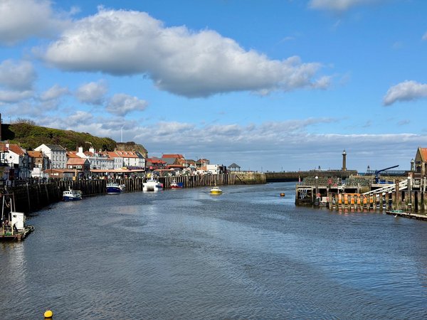 A small harbour with calm water is lined with fishing boats and a mix of white and grey buildings with red-tiled roofs along the left side. The quayside is reinforced with wooden pilings, and a few people can be seen walking near the waterfront. On the right, the harbour opens toward the sea, with a pier extending outward, marked by a tall lighthouse near the horizon. Several small boats, including a yellow one in the centre, are moored or slowly moving in the water. The sky is mostly clear with scattered clouds, and the scene is brightly lit under daylight, giving a clear view of the coastal landscape.